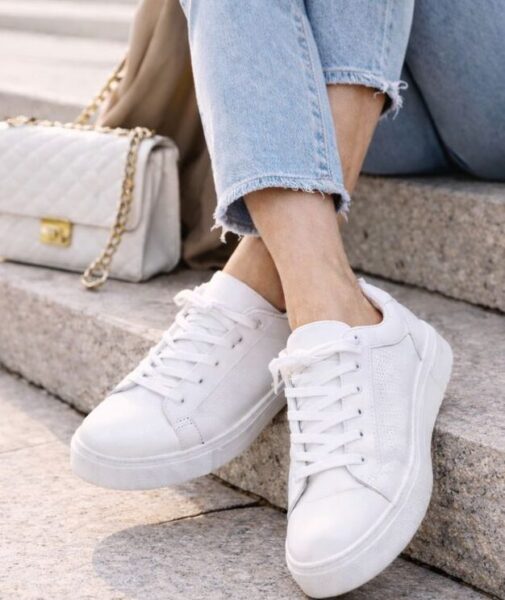 Weiße Sneakers - Eine Frau über 50 sitzt auf einer Treppe. Sie trägt weiße Sneakers zu einer Jeans und einem Trenchcoat kombiniert.