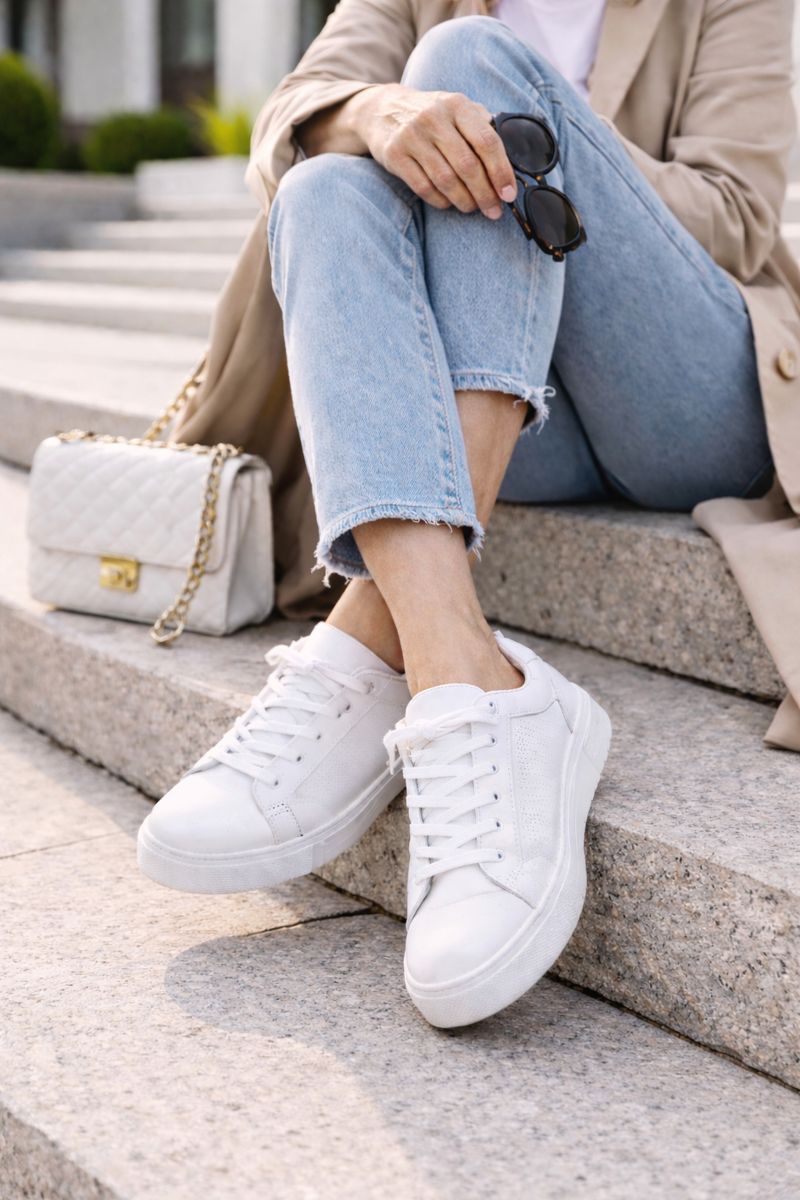 Eine Frau über 50 sitzt auf einer Treppe. Sie trägt weiße Sneakers zu einer Jeans und einem Trenchcoat kombiniert.